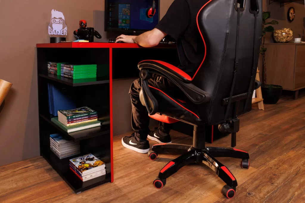 Mesa gamer Madesa, decorada com computador, mouse, livros e itens geek. À frente, cadeira gamer nas cores preto e vermelho.