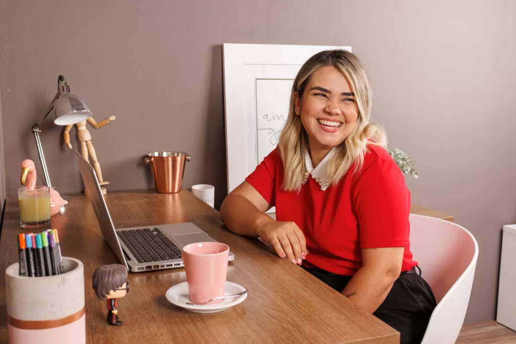 Uma mulher loira está sentada em uma cadeira que está na frente de uma escrivaninha Madesa. Ela está sorrindo e olhando para a direita e, sobre a escrivaninha à sua frente, estão dispostos um notebook, uma xícara de café, canetas, objetos decorativos e uma luminária.