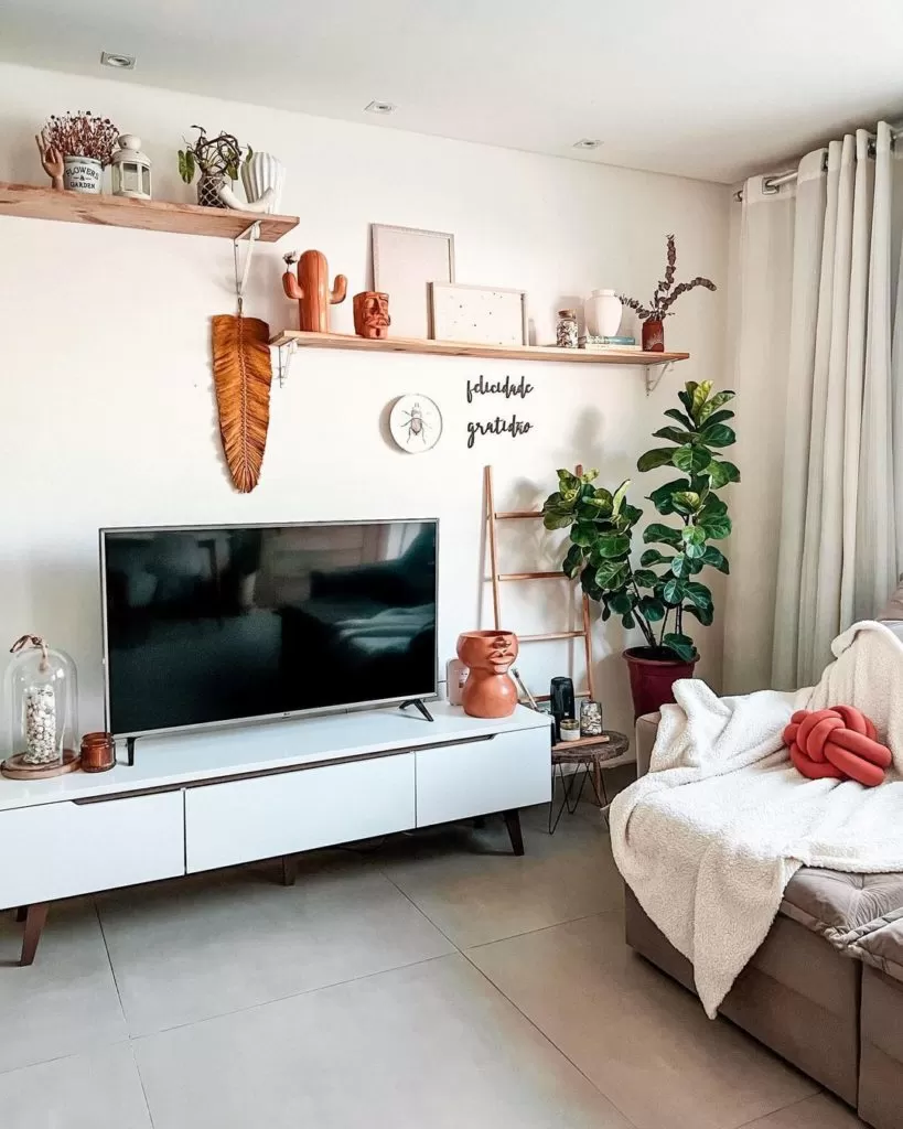 em uma sala com paredes claras estão dispostos diversos objetos decorativos em tons terrosos, além de plantas e uma televisão.