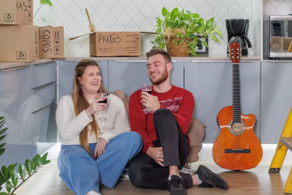 um casal está sentado ao chão de uma cozinha. Ao fundo, armários de uma cozinha Madesa contém caixas de mudança, eletrodomésticos e um vaso com plantas. O casal bebe vinho e, ao seu lado, está um violão. 