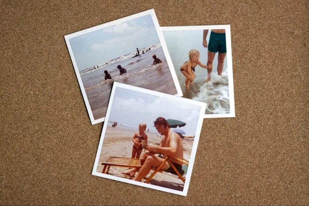  três fotografias polaroid estão dispostas sobre uma superfície marrom. Elas contém fotos de uma família aproveitando o dia na praia, como pais brincando com seus filhos na areia e no mar.