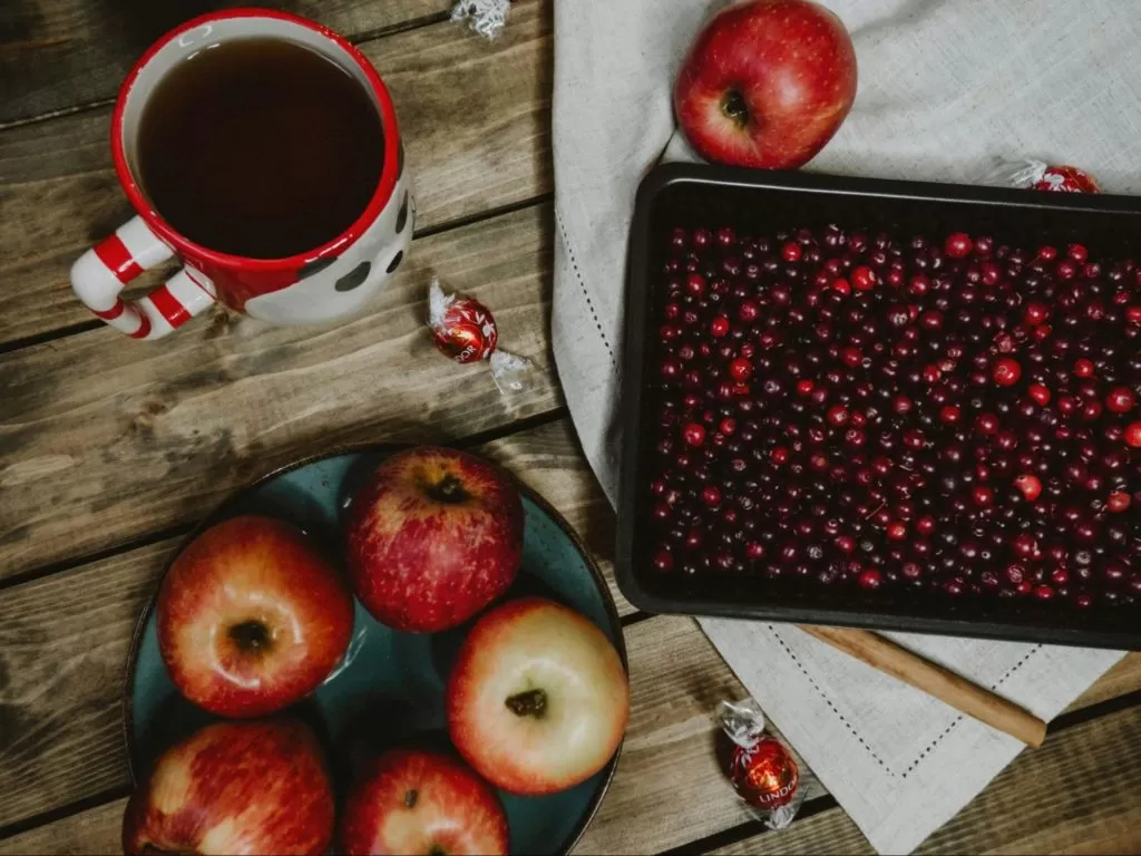 maçãs e cerejas dispostas sobre refratários. Ao lado, xícara com temática de natal e mesa de madeira abaixo