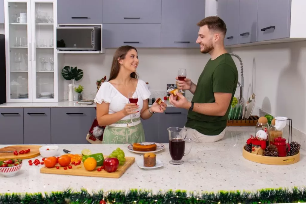 alt text: casal veste verde e conversa e ri em cozinha Madesa. Em cima do balcão estão decorações festivas verdes e vermelhas, suco de uva e frutas