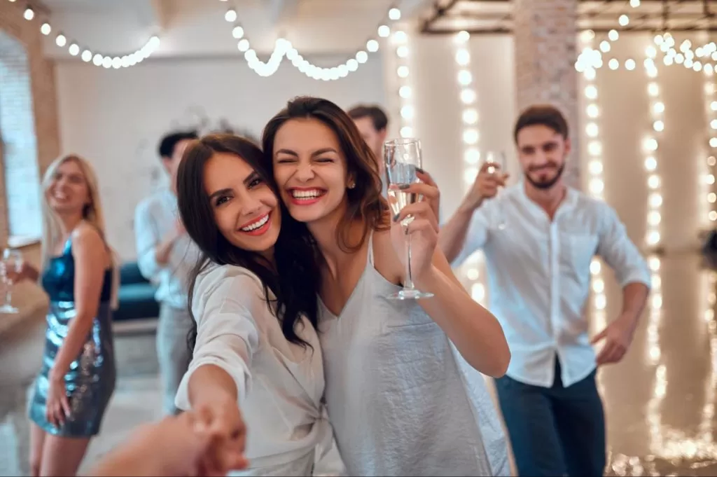 pessoas em festa de ano novo com luzes amarelas e taças na mão, vestem branco e paete e sorriem para a câmera, como se tirassem uma selfie