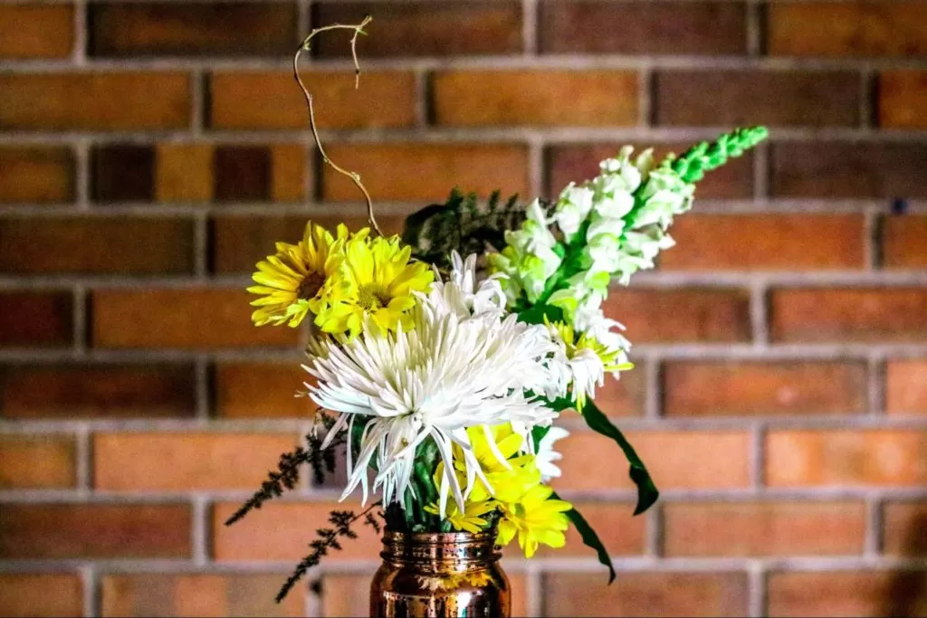 um vaso de metal acobreado com arranjo de flores branca e amarelas, folhagens e galhos secos está em foco, em frente a parede de tijolos à vista