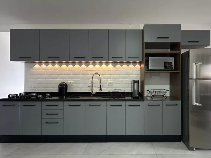 Cozinha moderna com armários em tom cinza e bancada de granito preto. A iluminação é proporcionada por lâmpadas embutidas no teto, criando um ambiente bem iluminado. A janela permite a entrada de luz natural, e há detalhes decorativos como plantas e utensílios organizados na bancada.