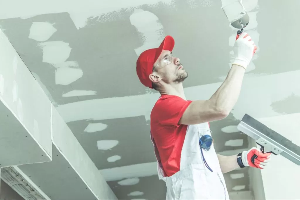 homem veste roupas vermelhas e macacão branco enquanto passa materiais de reforma na superfície de um teto de gesso