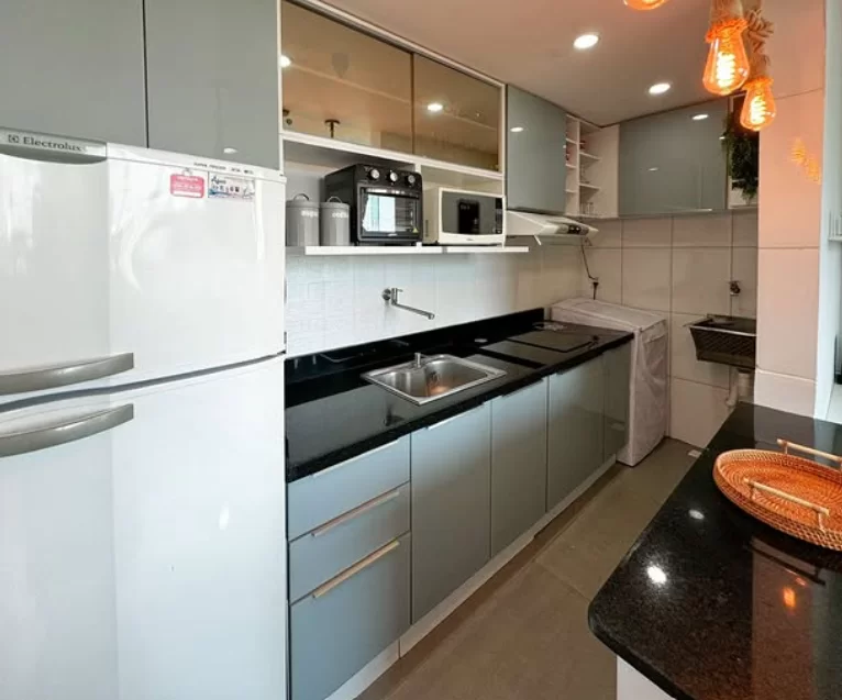 :Cozinha moderna com armários cinzas e bancada de granito preto. À esquerda, há um refrigerador branco. Na bancada, uma pia de aço inoxidável está visível. Acima da pia, um micro-ondas e uma cafeteira estão dispostos em prateleiras. À direita, uma lavadora de roupa está coberta com um tecido, e um tanque de lavar está disponível. Luminárias pendentes em formato de lâmpada iluminam o ambiente. Uma cesta decorativa está presente na borda da bancada.