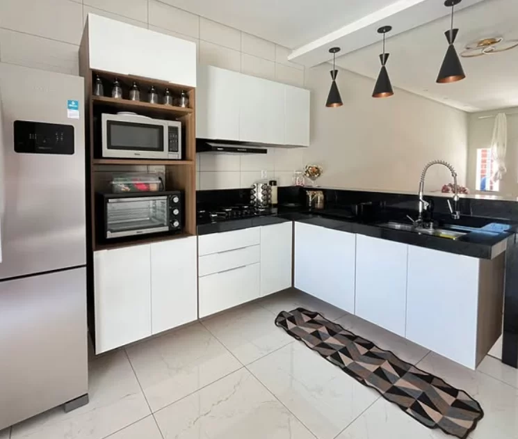 Cozinha moderna com armários brancos e detalhes em madeira. À esquerda, há um refrigerador prateado. Um armário em formato vertical contém um micro-ondas e um forno elétrico. O fogão a gás está ao lado, com uma coifa acima. A bancada é de granito preto, com uma pia e torneira. Três luminárias pendentes iluminam a área. Um tapete decorativo com padrões geométricos está no chão.