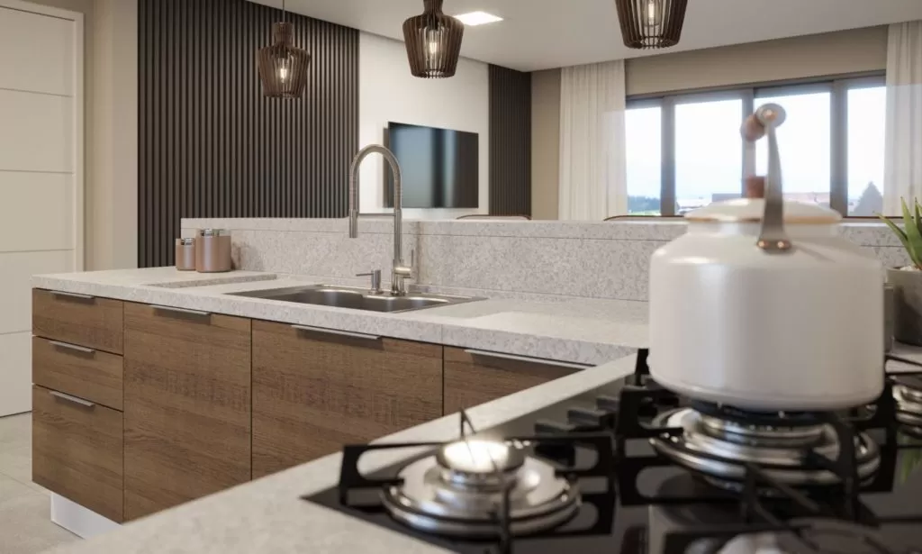 Cozinha moderna com armários de madeira e uma bancada clara. A bancada possui uma pia de aço inoxidável e um fogão a gás com queimadores. Em primeiro plano, há uma chaleira branca. Ao fundo, há luminárias pendentes em um design de lâmpada de vime, uma TV na parede, e amplas janelas com cortinas brancas que permitem a entrada de luz natural. Uma planta verde em um vaso cinza está visível na bancada.