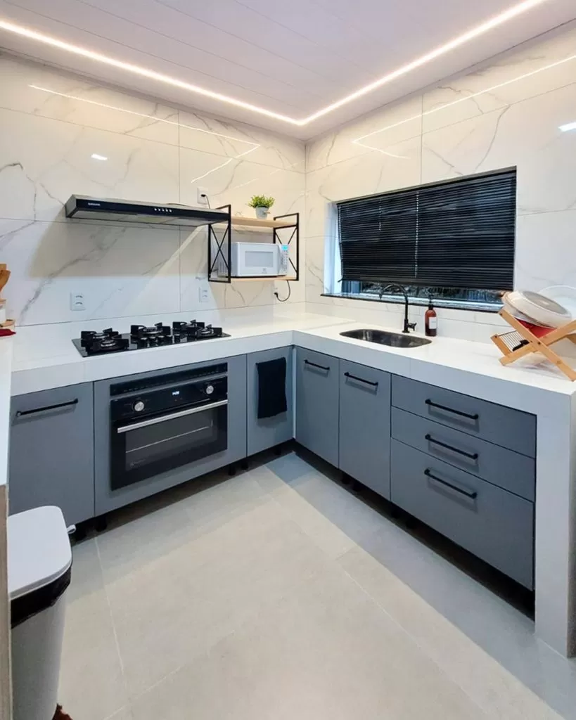 Cozinha com armários cinzas, bancada branca, cooktop de quatro bocas, forno embutido, uma janela com persiana preta, e uma prateleira com micro-ondas.