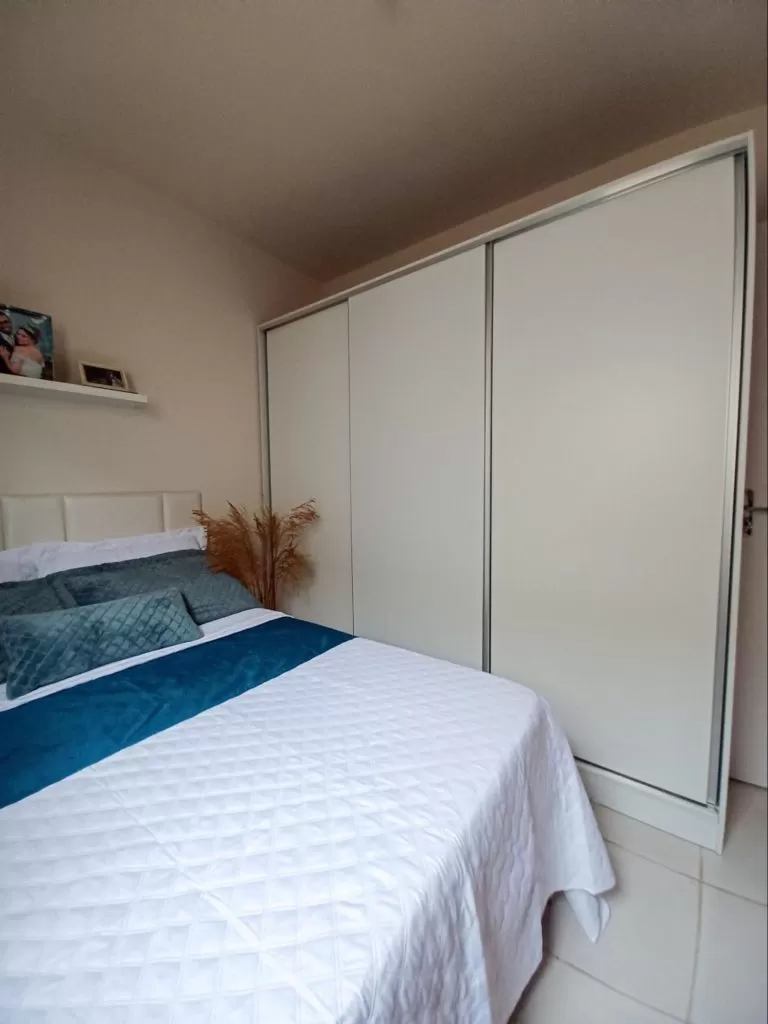 Guarda-roupa branco 100% MDF, com 3 portas de correr, ao lado de uma cama de casal com colcha branca e detalhes em tons de azul.
