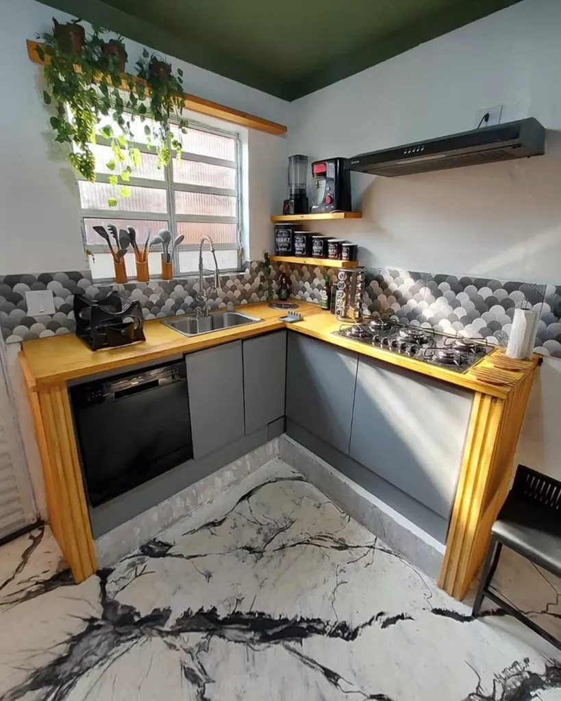 Cozinha de canto com móveis de madeira natural na bancada, armários de cor cinza com acabamento fosco, uma janela ao fundo com cortina branca, utensílios de cozinha na bancada e prateleiras com objetos decorativos e eletrodomésticos.