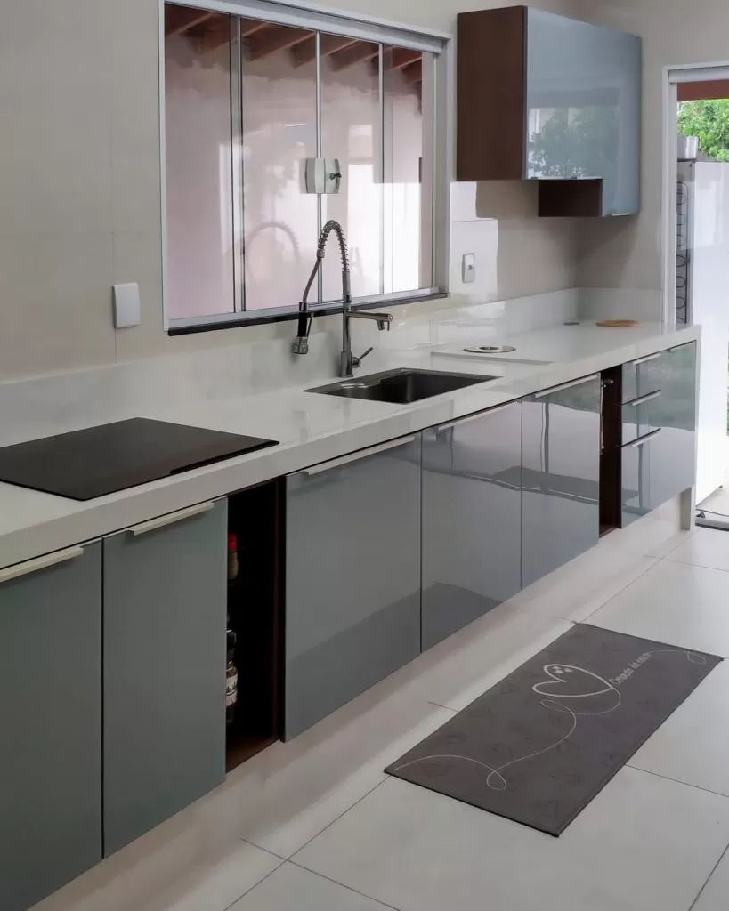 Cozinha com balcão de granito branco, armários de cor cinza com acabamento brilhante, portas espelhadas e uma janela ao fundo.