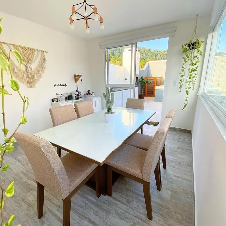 Mesa de jantar branca retangular com seis cadeiras bege, plantas em vasos no centro e ao redor, janelas grandes permitindo entrada de luz natural, e decoração moderna no ambiente.