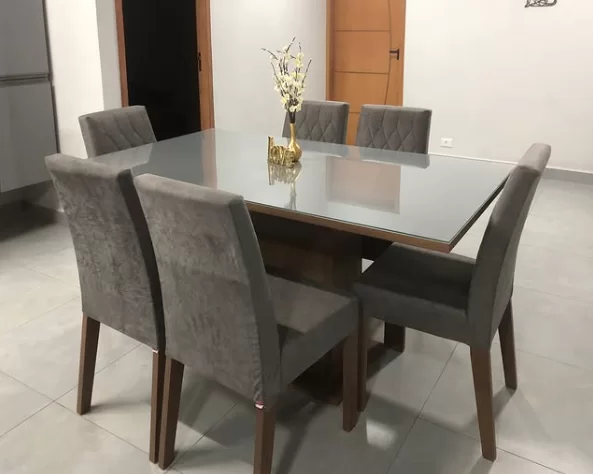 Mesa de jantar retangular com tampo de vidro e seis cadeiras cinza, decorada com um vaso dourado com flores brancas, em um ambiente com piso claro e paredes neutras.
