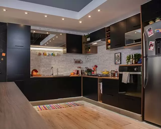Cozinha com armários pretos e detalhes em madeira, pia com torneira, parede de azulejo branco e iluminação embutida no teto. Há uma geladeira prateada com fotos e recados, e utensílios de cozinha coloridos na bancada. O piso é de madeira clara e há um tapete colorido na frente da bancada.