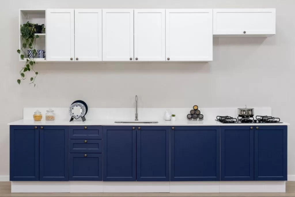 Balcão de cozinha com armários azuis na parte inferior, com três gavetas ao centro, e armários brancos na parte superior. No balcão, há pratos decorativos, utensílios de cozinha, uma pia com torneira, e uma panela no fogão ao lado.