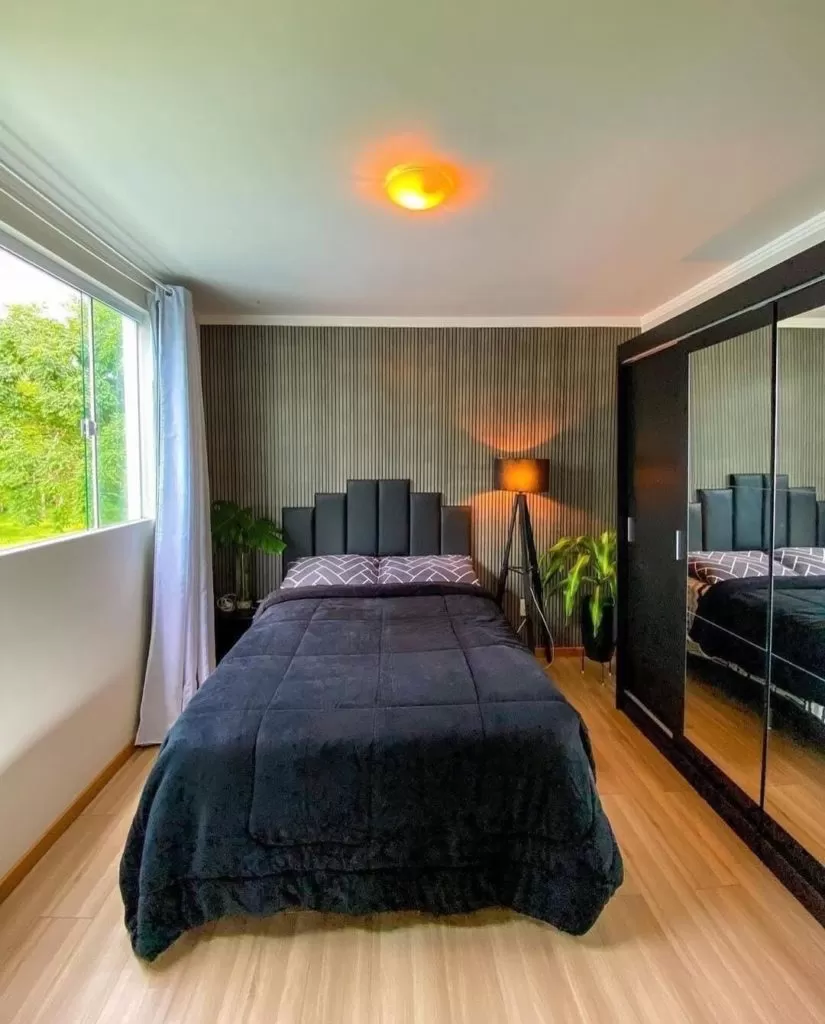 Quarto moderno com decoração em tons escuros. A cama com edredom e cabeceira pretos está centralizada em frente a uma parede com painel ripado cinza. À direita, um guarda-roupa preto com porta de espelho, e à esquerda, uma grande janela com vista para a natureza. O piso é de madeira clara.