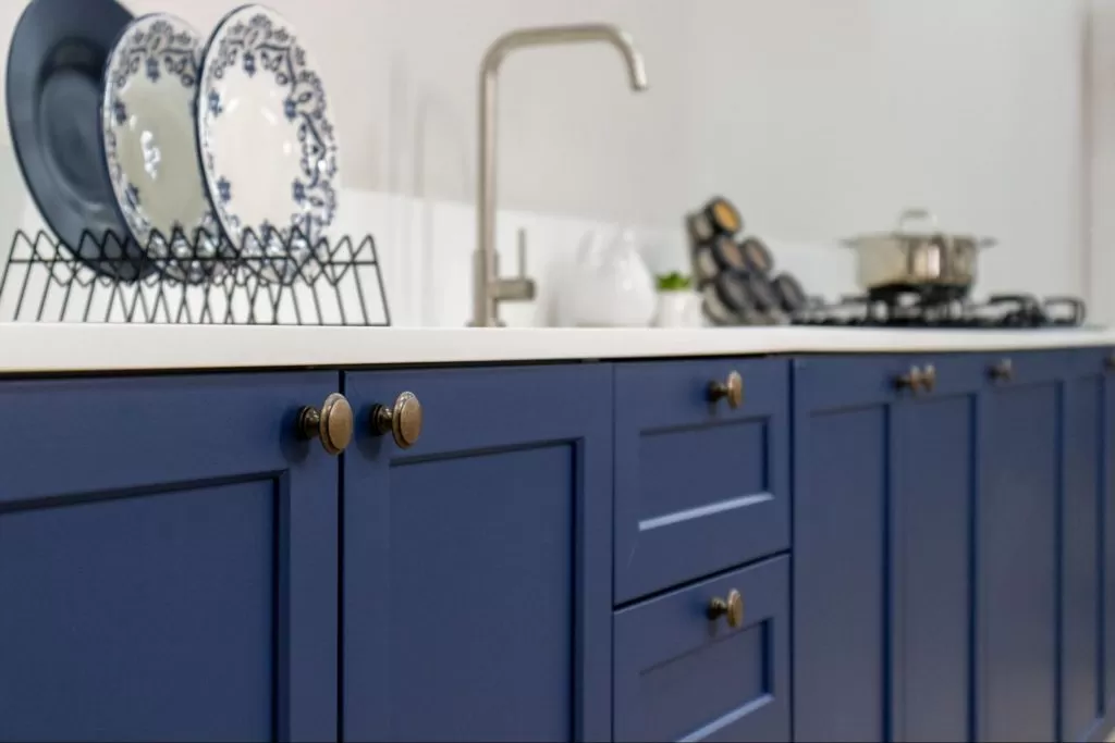 Armário de cozinha azul com portas e gavetas, com detalhes em maçanetas douradas, e pia com torneira ao fundo. Ao lado, utensílios de cozinha, como pratos decorativos brancos com detalhes azuis em um escorredor e uma panela no fogão.