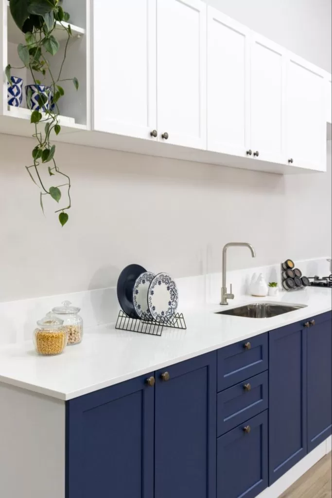 Balcão de cozinha com armários azuis na parte inferior, com três gavetas ao centro, e armários brancos na parte superior. No balcão, há um escorredor com pratos decorativos e utensílios de cozinha, além de uma pia com torneira, uma panela no fogão e alguns itens decorativos.