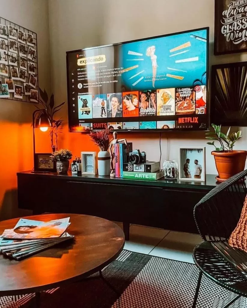 Sala de estar aconchegante com uma TV na parede, exibindo a tela da Netflix. Abaixo, um rack preto está repleto de decorações como livros, uma câmera vintage e porta-retratos. A parede atrás da TV é iluminada por uma luz laranja quente. Ao lado, há um mural de fotos e, no primeiro plano, uma mesa de centro e uma cadeira preta.
