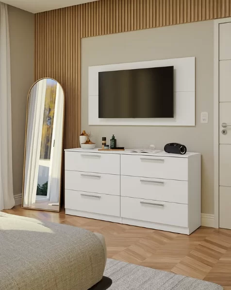 Quarto com uma cômoda branca de seis gavetas, uma televisão na parede acima e um espelho de corpo inteiro apoiado na parede ao lado. Sobre a cômoda, há objetos decorativos e eletrônicos.