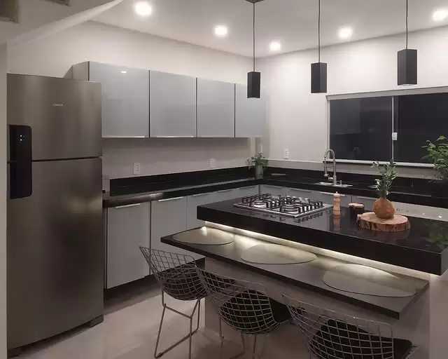 Cozinha com armários cinzas, bancadas de granito preto e uma ilha central com cooktop. A ilha possui uma bancada mais baixa para refeições, iluminada por uma fita de LED e acompanhada por três cadeiras de design aramado. Três pendentes pretos e uma geladeira de inox completam o ambiente.