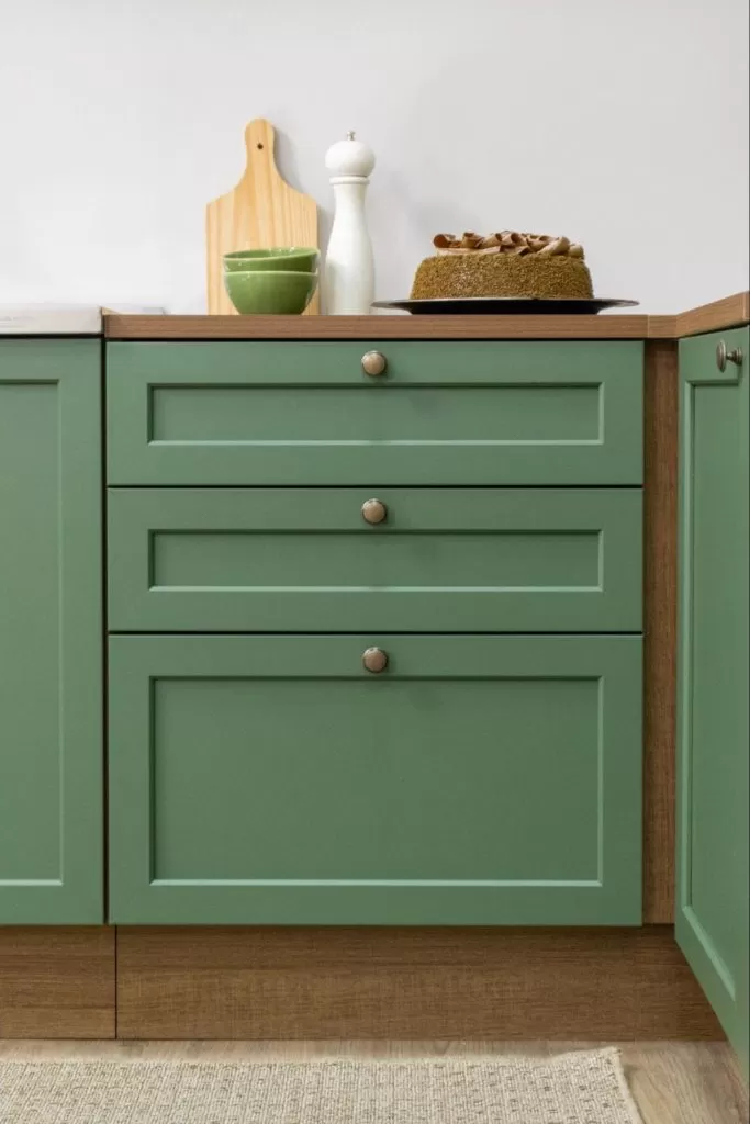 Parte de um armário de cozinha verde com três gavetas, com detalhes em botões metálicos, e uma bancada de madeira clara. No topo, há uma tábua de cortar de madeira, dois bowls verdes, um saleiro branco, um bolo decorado e uma panela no fogão ao lado.
