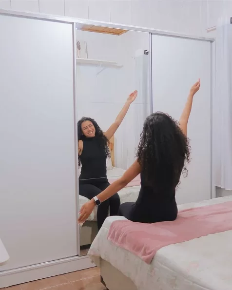 Uma mulher com cabelos cacheados, vestindo roupa preta, sentada na cama de frente para um espelho grande, com os braços levantados e sorrindo.