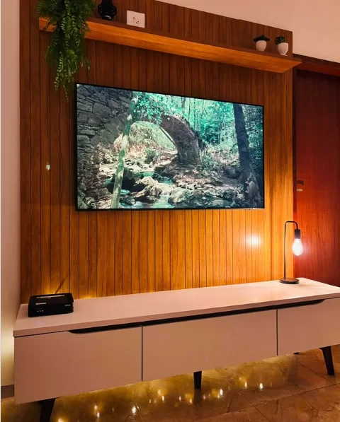 Sala de estar com um painel de madeira ripado na parede. Uma TV está montada no painel, acima de um rack branco de estilo moderno com pés escuros. Uma prateleira de madeira no topo exibe plantas, e uma fita de LED na base do rack ilumina o piso brilhante.
