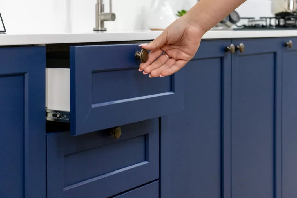 Cozinha Vik na cor azul com caixaria branca. O foco está nas gavetas do balcão de pia sendo abertas por uma mão feminina. Bancada de granito branca com uma torneira de inox ao fundo.
