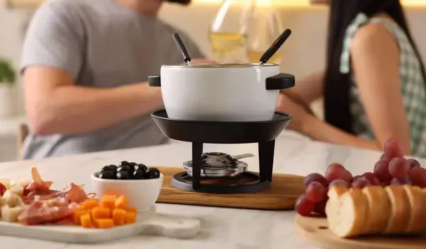 Uma panela de fondue branca sobre um suporte de aquecimento, com dois garfos de fondue inseridos, em uma mesa de jantar. Ao redor, há pratos com uvas, queijo, presunto, cenoura picada, e um pão cortado em fatias. Pessoas ao fundo conversando.