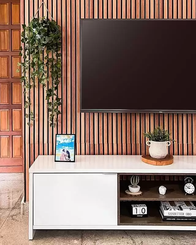 Interior de uma sala com painel de madeira na parede, uma televisão grande pendurada na parede, uma planta pendurada em um suporte na parede, uma planta em um vaso decorativo branco sobre uma bandeja de madeira, uma moldura com uma foto de casal e objetos decorativos na prateleira do móvel.