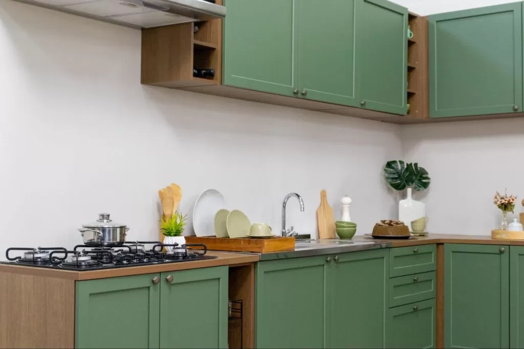 Cozinha com armários verdes e detalhes em madeira, pia com torneira, bancada com pratos, tigelas e utensílios decorativos, incluindo vasos com plantas e um bolo, em um ambiente bem organizado e decorado com elementos naturais.