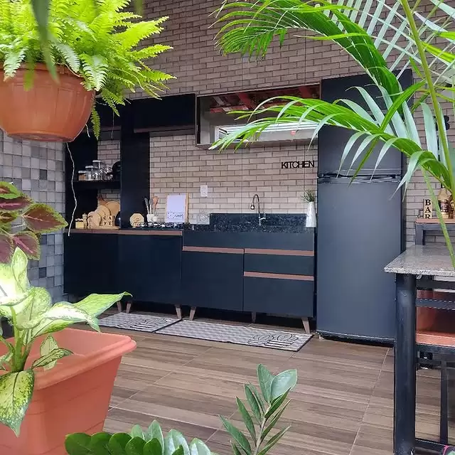 Cozinha externa com paredes de tijolos aparentes e armários pretos. Pia com bancada de granito, uma geladeira preta ao lado, decorada com plantas ao redor, incluindo uma planta pendurada em um vaso de barro e outras em vasos no chão.