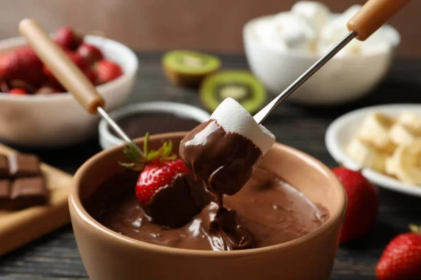 Uma panela de fondue de chocolate com marshmallow e morango sendo mergulhados na mistura. Ao fundo, há tigelas com frutas, chocolate e outros ingredientes para fondue, e uma tábua com pedaços de chocolate.