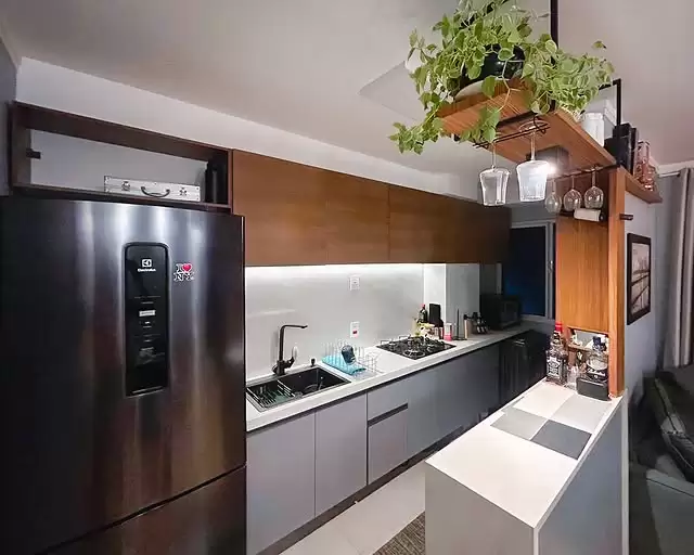 Cozinha com balcão branco na frente, armário aéreo em madeira acima da pia, prateleiras abertas de madeira com plantas, copos e utensílios. Ao lado, uma geladeira inox e uma parede de fundo branca.