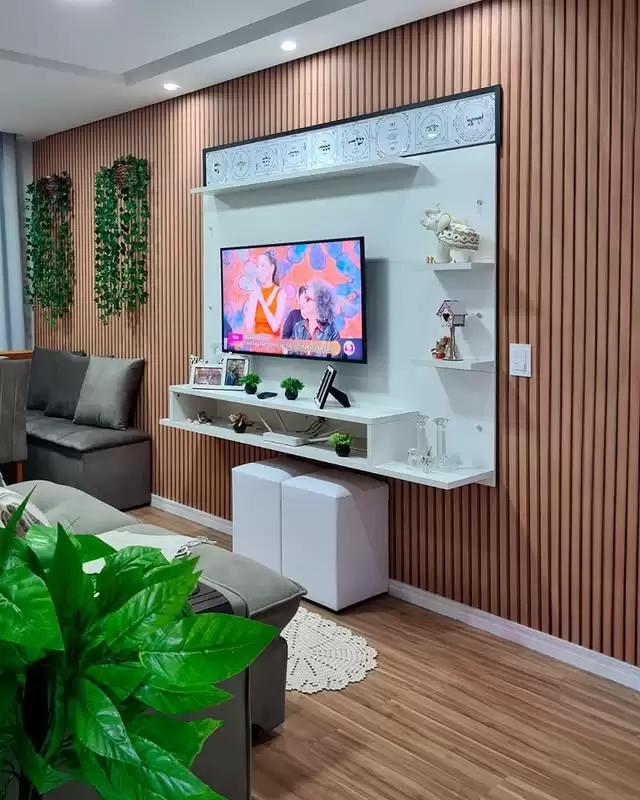 Sala de estar com parede de painéis de madeira, televisão de tela plana instalada na parede, prateleiras brancas decoradas com plantas, objetos decorativos, sofá cinza ao lado, plantas penduradas na parede e uma planta grande no canto inferior esquerdo, além de uma pequena área com tapete de crochê.