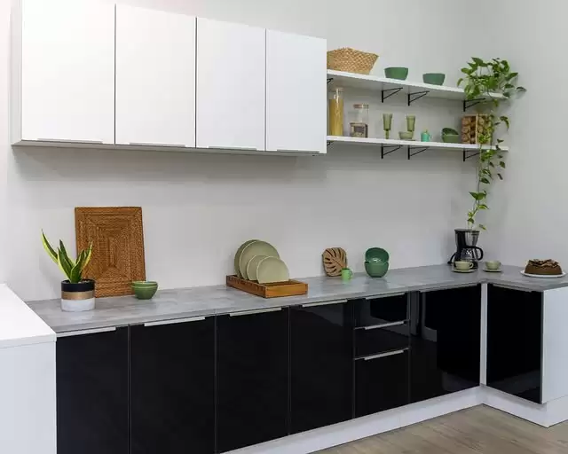 Cozinha com armários pretos na parte inferior e armários brancos na parte superior. Prateleiras brancas com vasos verdes, pratos e objetos decorativos. Na bancada de mármore cinza-claro, há uma planta em vaso, pratos empilhados, vasos decorativos, uma cafeteira e uma torta.