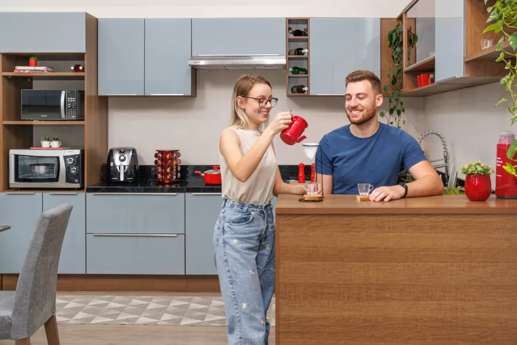 Duas pessoas brancas em frente a uma cozinha cinza, sobre uma bancada em tom amadeirado em cozinha organizada na rotina.