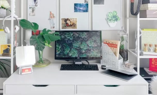 Escritório em casa equipado com escrivaninha branca e um computador preto. Na tela do monitor há imagens de plantas.