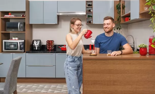 Duas pessoas brancas em frente a uma cozinha cinza, sobre uma bancada em tom amadeirado em cozinha organizada na rotina.