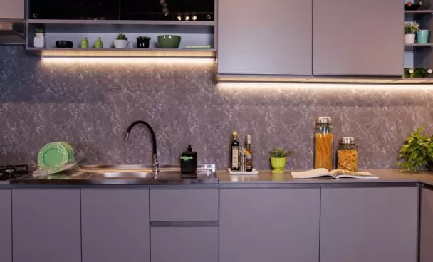 Ambiente com uma pia de cozinha e bancada cinza. A parte superior exibe prateleiras com plantas e utensílios. Louças verdes estão dispostas ao lado da pia.