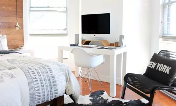 Quarto pequeno em tons claros. Na imagem podemos ver uma parte da cama, uma escrivaninha com computador e uma cadeira com uma almofada preta.