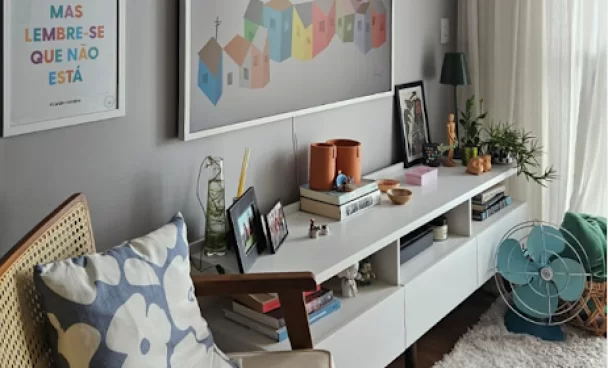 Sala de estar com sofá de madeira e almofada com estampa floral. Uma mesa branca exibe livros, uma caneca, plantas e quadros com fotos.