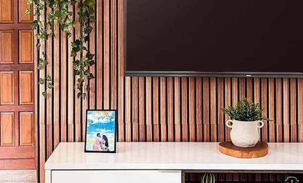 Interior de uma sala com painel de madeira na parede, uma televisão grande pendurada na parede, uma planta pendurada em um suporte na parede, uma planta em um vaso decorativo branco sobre uma bandeja de madeira, uma moldura com uma foto de casal e objetos decorativos na prateleira do móvel.