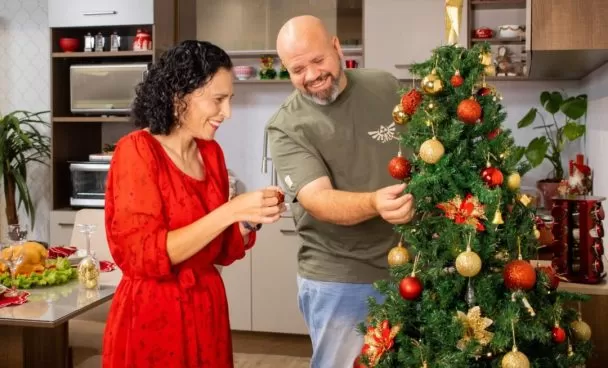 Casal de adultos, decorando juntos uma árvore de Natal, sorrindo. Ao fundo, uma cozinha Madesa e uma mesa de jantar, ambos decorados.
