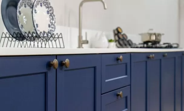 Armário de cozinha azul com portas e gavetas, com detalhes em maçanetas douradas, e pia com torneira ao fundo. Ao lado, utensílios de cozinha, como pratos decorativos brancos com detalhes azuis em um escorredor e uma panela no fogão.