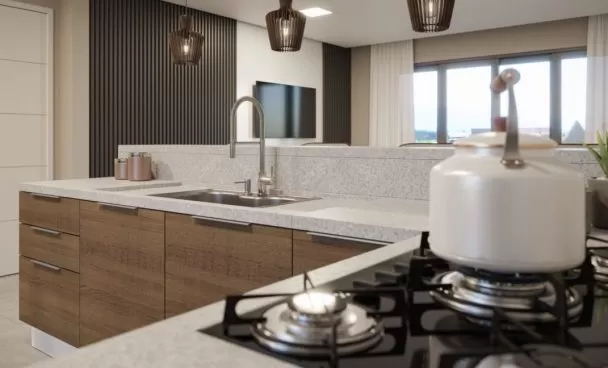 Cozinha moderna com armários de madeira e uma bancada clara. A bancada possui uma pia de aço inoxidável e um fogão a gás com queimadores. Em primeiro plano, há uma chaleira branca. Ao fundo, há luminárias pendentes em um design de lâmpada de vime, uma TV na parede, e amplas janelas com cortinas brancas que permitem a entrada de luz natural. Uma planta verde em um vaso cinza está visível na bancada.